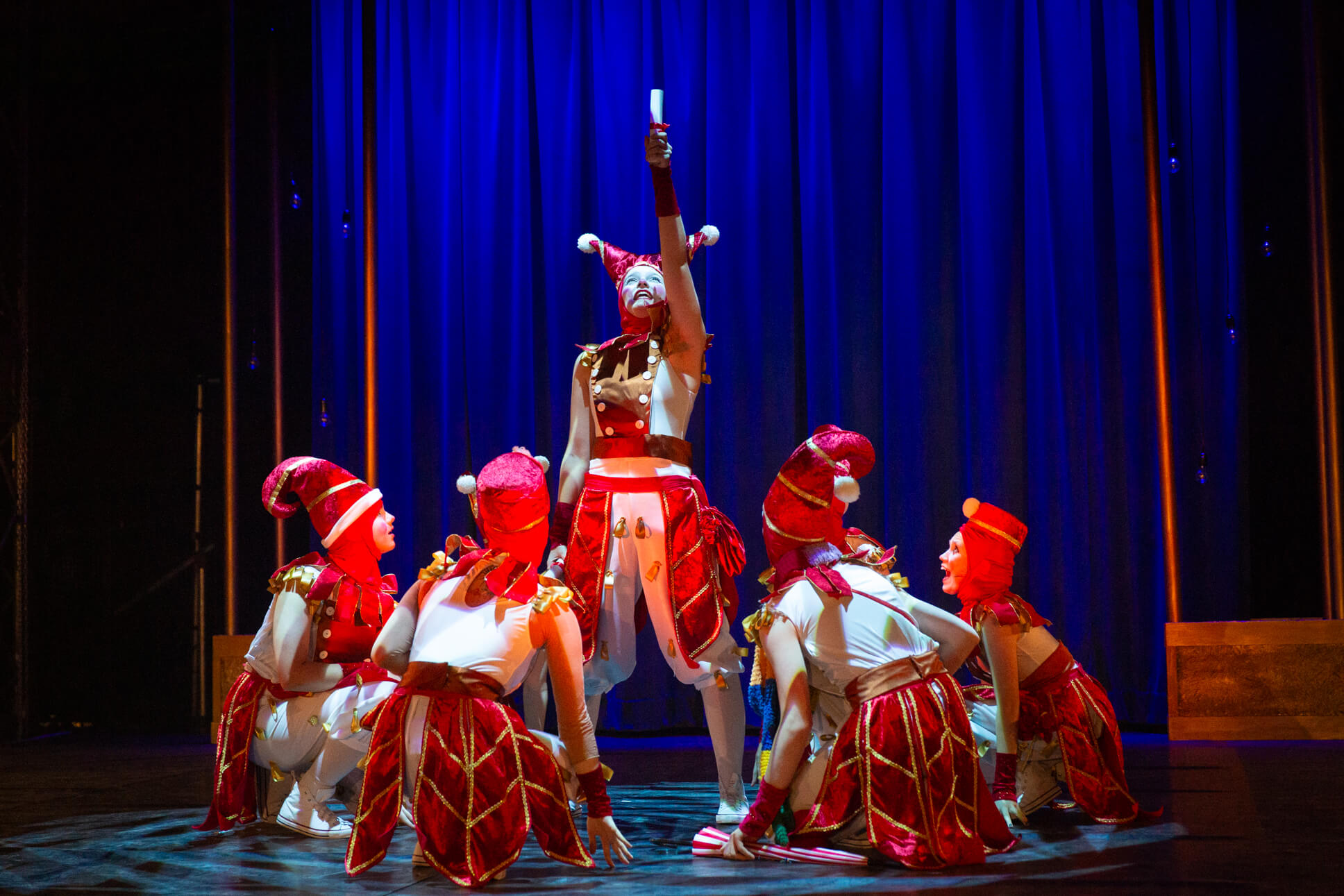 A dynamic dance sequence from a stage performance where performers in red and white jester costumes form a circle, lifting one performer who reaches skyward against a deep blue curtain backdrop