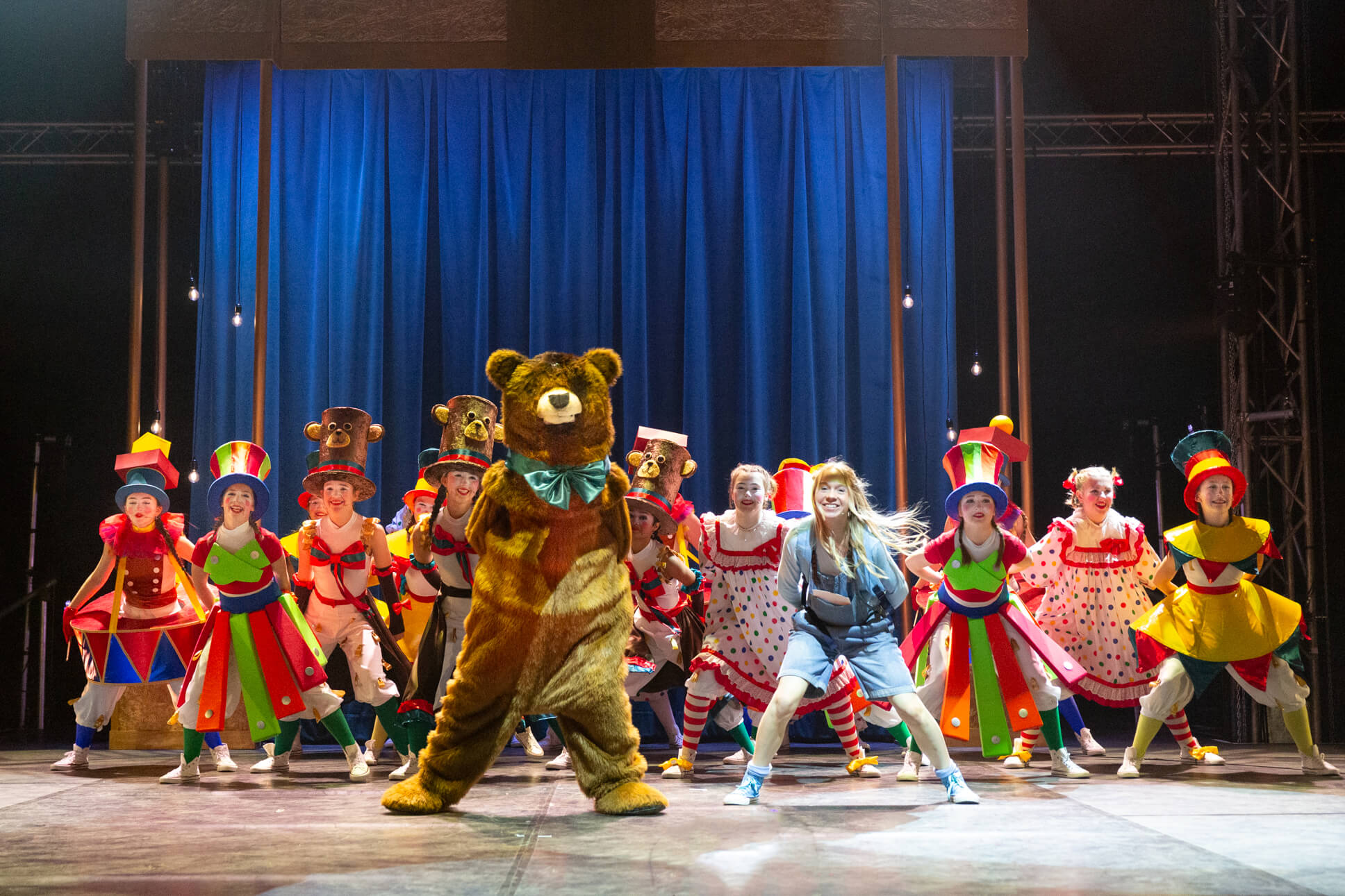 An energetic dance number featuring performers in colorful carnival costumes and monkey-topped hats surrounding a large brown teddy bear character center stage, with a royal blue curtain backdrop