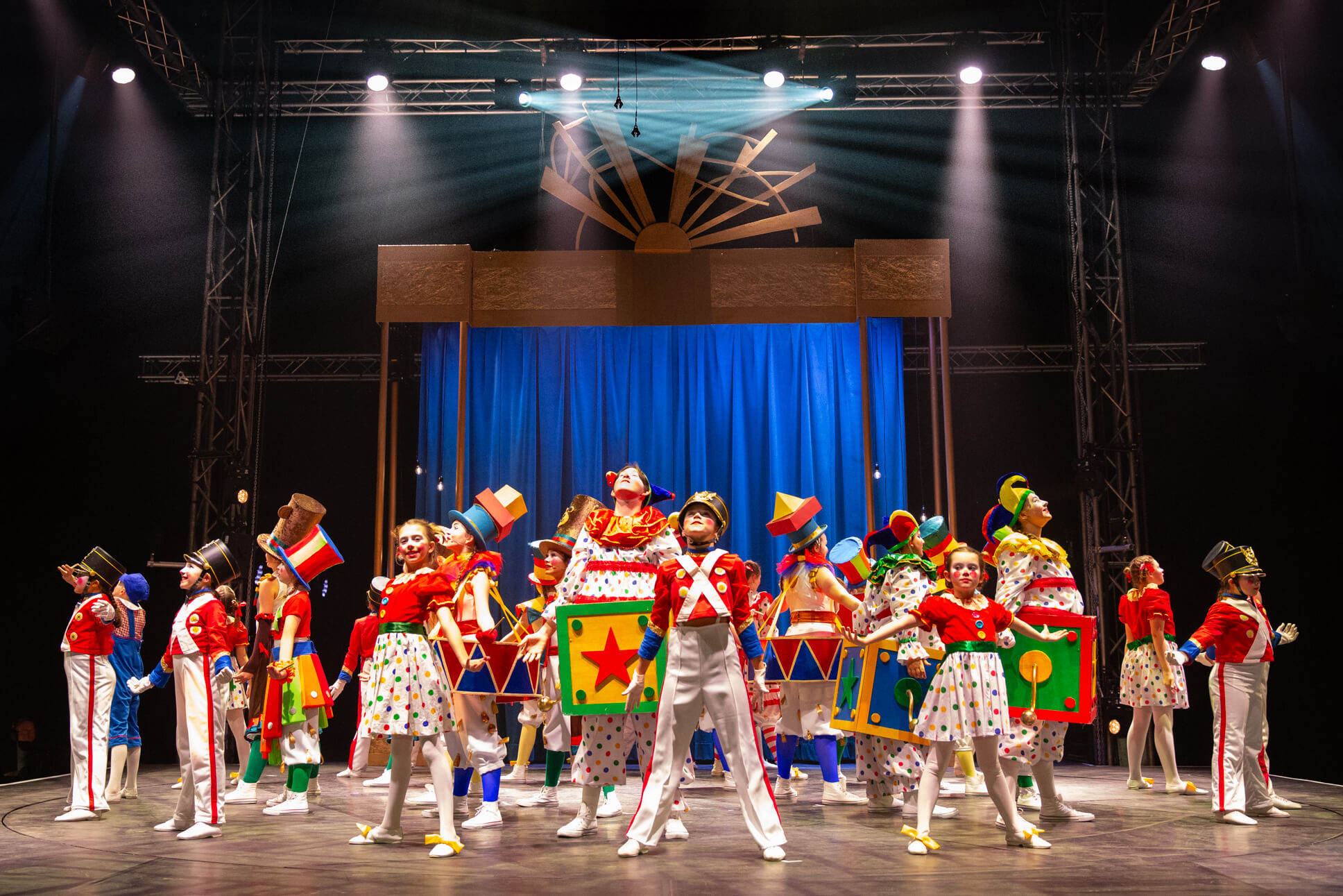 A theatrical ensemble scene with performers in toy soldier and polka-dot dress costumes arranged in formation, featuring decorative toy blocks and a stylized sun set piece above the stage