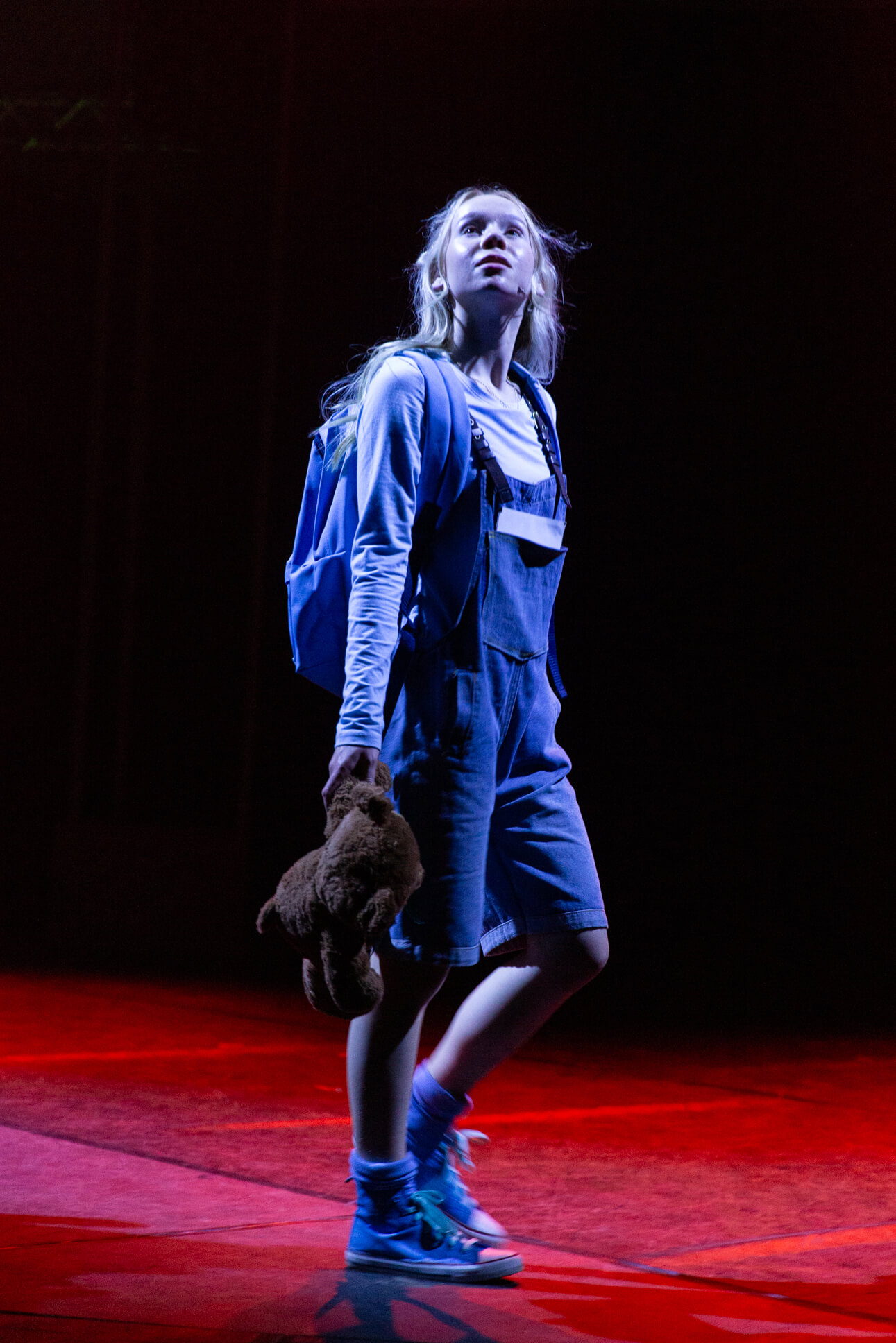 A dramatic solo scene of a performer in denim overalls and a blue backpack, holding a teddy bear while standing on a red-lit stage, creating a striking silhouette against the dark background