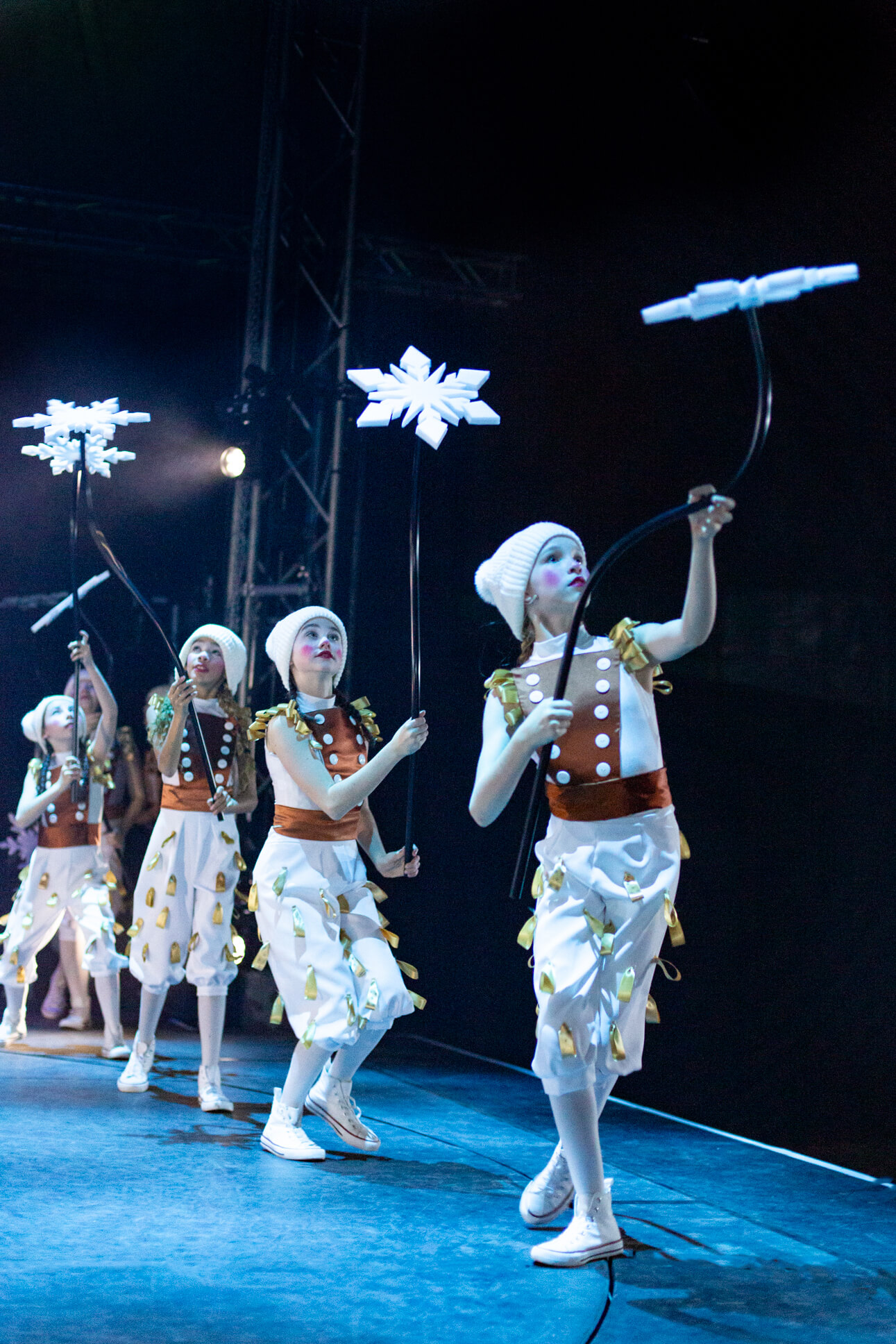 A line of performers in white knit caps and white costumes with copper-colored sashes hold up illuminated snowflake props against a dark stage. Their costumes feature decorative gold ribbons, and they wear white sneakers as they perform in synchronized poses under blue lighting