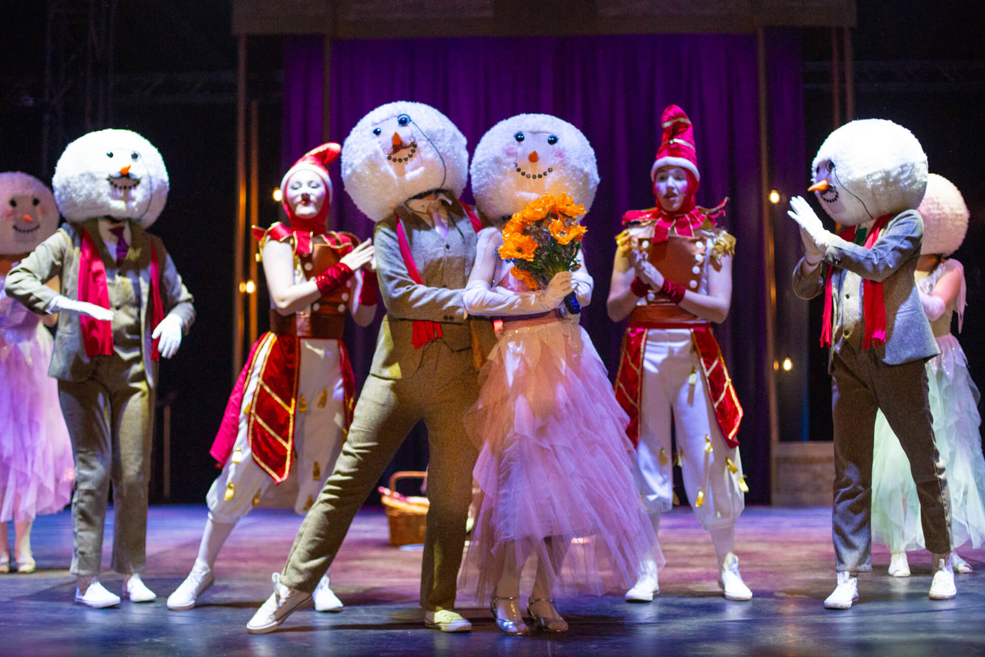 A romantic scene between two performers in snowman costumes - one in a suit and one in a tulle dress holding orange flowers - surrounded by other snowman characters and performers in red and white costumes. The snowmen wear large white spherical head pieces with carrot noses and beaded smiles